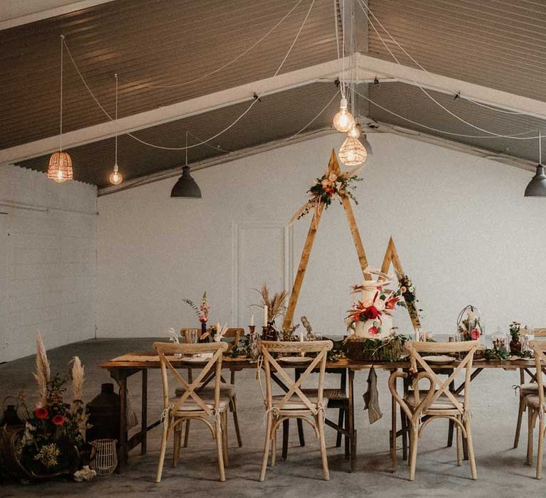 One small and one large wooden triangular wedding arch with pampas grass, foliage, and garden rose arrangements attached, rustic wedding decorations, hanging lampshades, and boho rustic wedding tablescape at The Barn at Avington