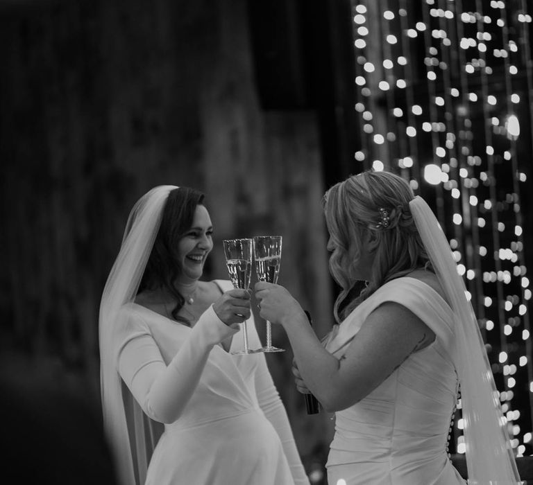 Bride in long sleeve deep v wedding dress and church length veil and bride in satin short sleeve deep v wedding dress and church length veil having a cheers of champagne at LGBTQIA wedding at Hidden River Cabins 