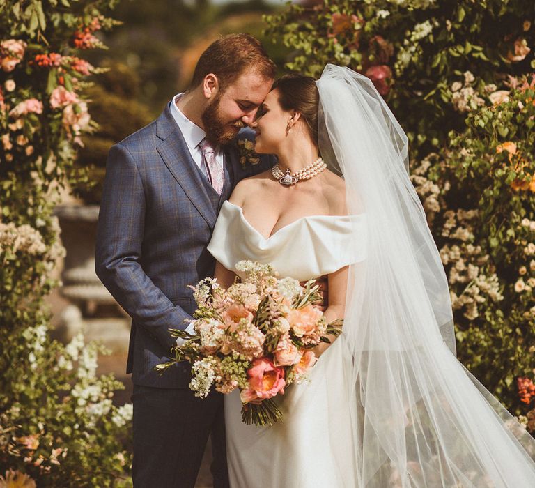 Bride wearing pearl choker necklace by Vivienne Westwood kissing groom in blue suit for classic wedding 