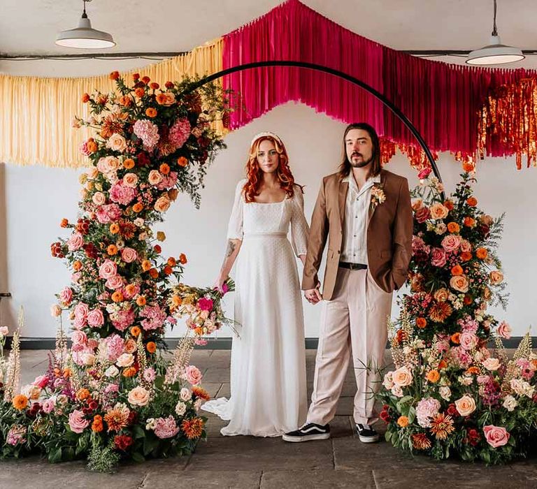 Groom in brown blazer, white shirt, grey suit trousers and vans shoes holding hands with bride in three quarter length puff sheer sleeve wedding dress with delicate polka dot overlay and square neck standing underneath circular floral arch with vibrant flowers and foliage attached and pink, red, yellow and metallic wedding streamers behind at Northern Monk Refectory Leeds