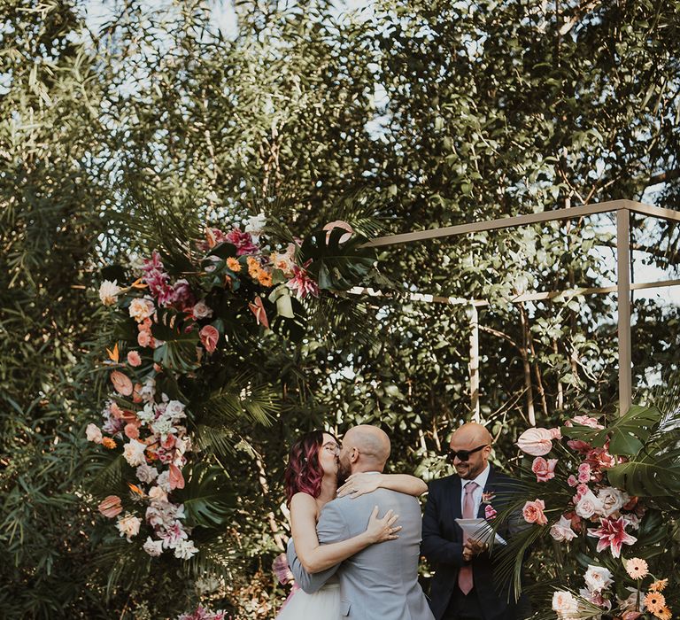 bride in a layered wedding dress kissing her groom in a grey suit exchanging vows at their outdoor Italian villa wedding venue with tropical flower arch