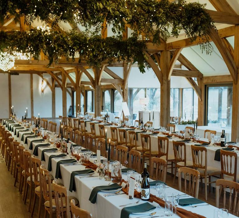 Classic wedding tablescape with white tablecloth, burnt orange wedding tablerunner, forest green wedding napkins, forest green tapered candles and bottles of wine with suspended foliage and fairy lights decor at wedding