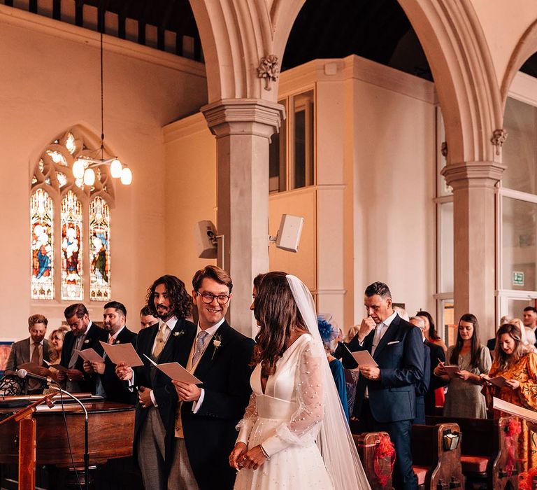 Bride in long sleeve pearl wedding dress by Sassi Holford with groom in morning suit reading out their wedding vows at church ceremony 