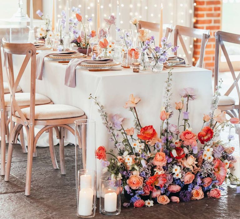 Peach and lilac wedding tablescape with spring flower centrepieces, lilac napkins, tinted glassware and gold cutlery and crockery on white tablecloth with flower arrangement with peach garden roses, white peonies, lilac sweet peas, bluebells and wildflowers at Rackleys Barn