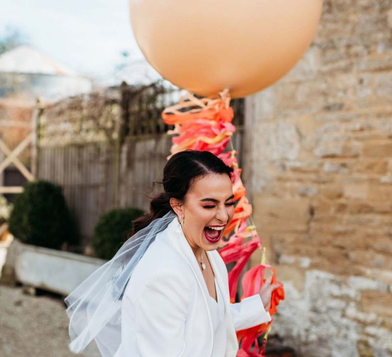 Bride in white bridal suit with closed toe bright green wedding heels and short bridal veil laughing outside Merriscourt wedding venue holding large balloon with colourful wedding streamers attached