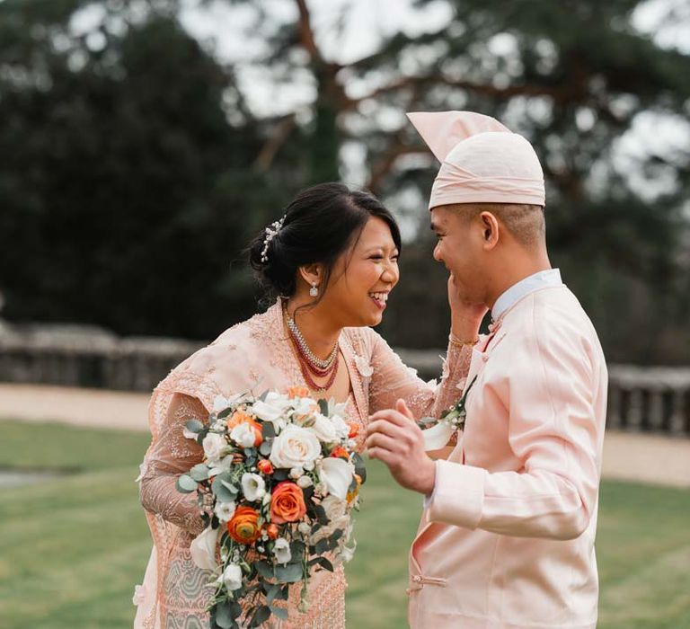 Groom in baby pink htaingmathein laughing with bride in floral 3D applique pink and rose gold htamane holding garden rose, poppy and foliage bridal bouquet at Burmese wedding