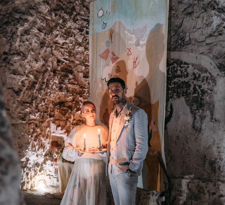 Groom in baby blue linen suit, personalised embroidered linen shirt and garden rose and pampas grass boutonniere standing with bride in sheer overlay wedding dress and fluffy bridal coverup holding candles up  next to large wedding fabric banner at Margate Caves 