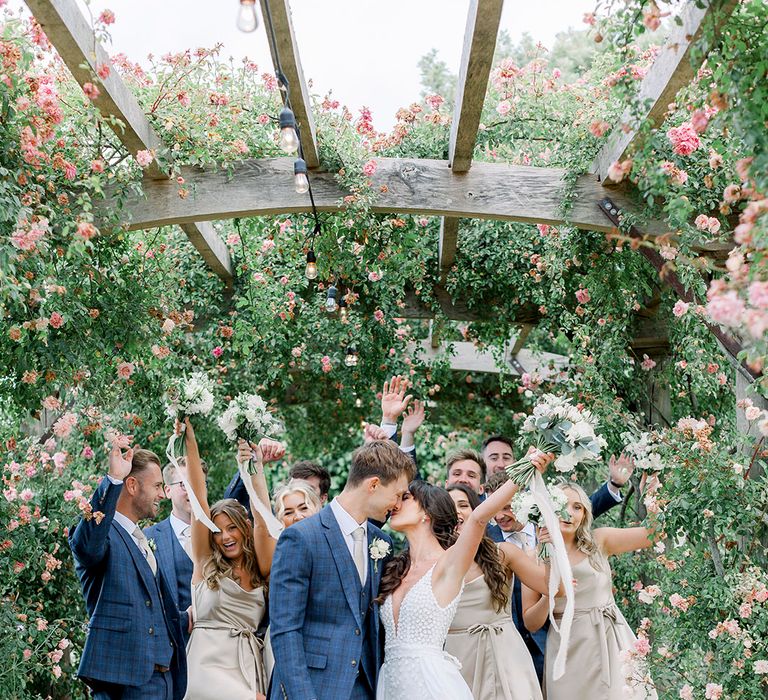 wedding party in navy suits and champagne gold bridesmaid dresses cheering at groom in a check suit kisses his bride in an appliqué wedding dress