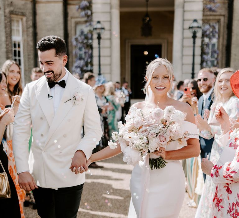 Bride carrying pastel pink and white wedding bouquet walking with groom in white suit jacket and black bow tie 