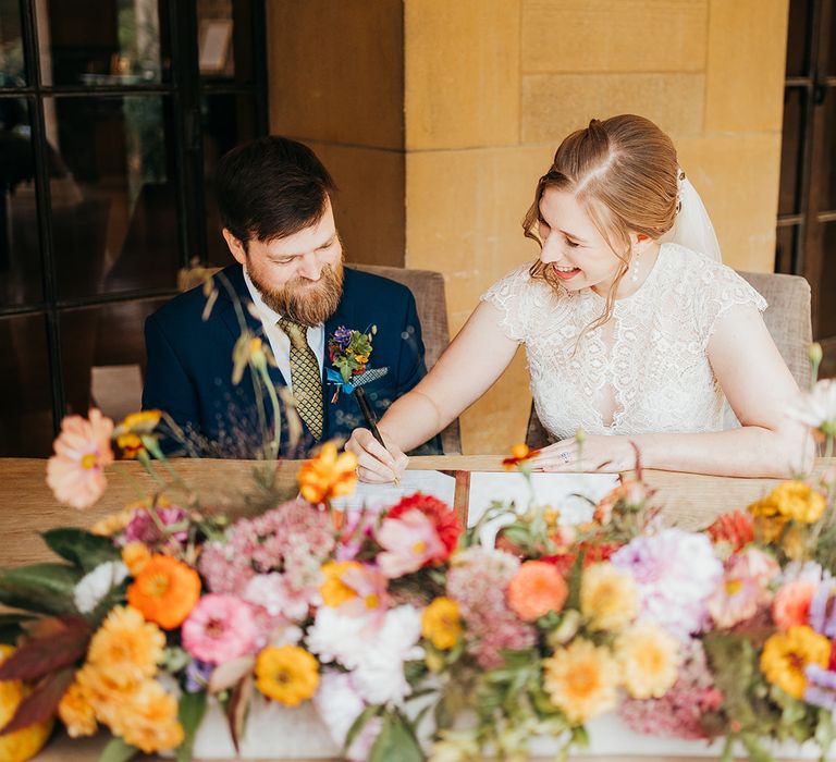 The bride and groom sit at a table decorated with seasonal autumn wedding flowers to sign the register 