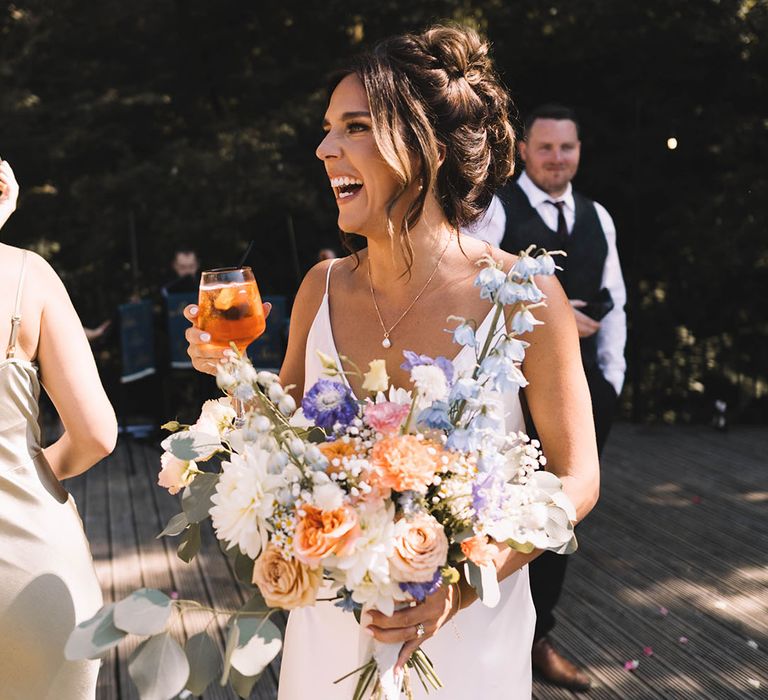 Bride wearing satin slip wedding dress holding pastel wedding bouquet drinking champagne 