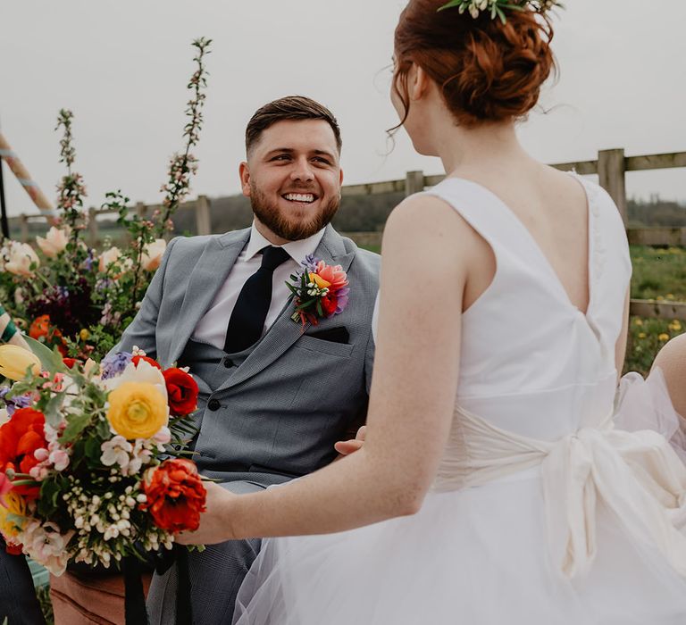 The groom lounges in a grey three piece suit facing the bride with her hair in an updo at their festival wedding 