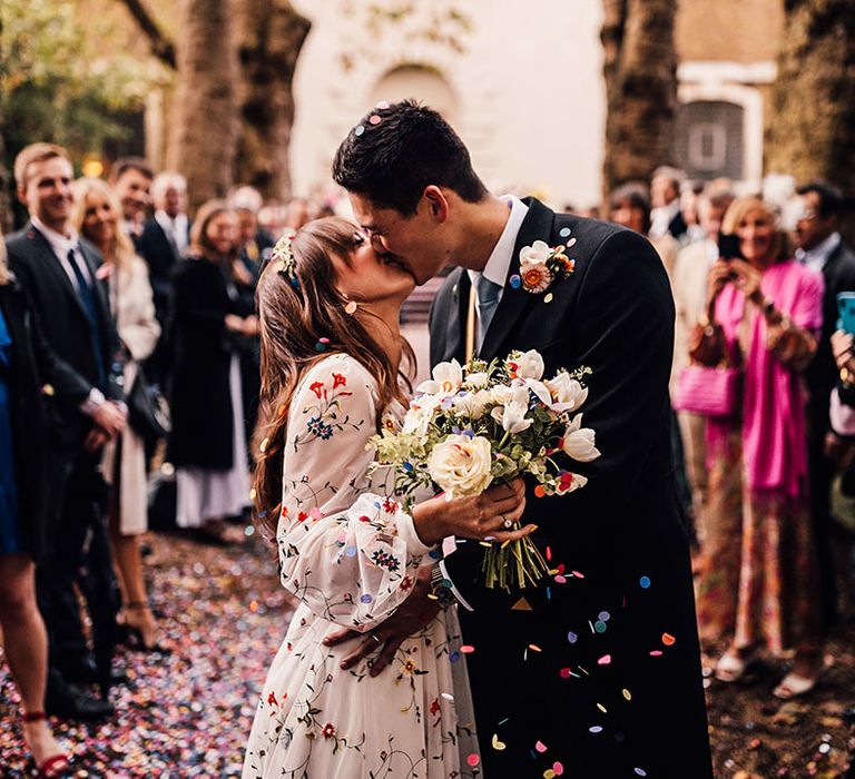 Bride in sheer long sleeve wedding dress kissing the groom as confetti is thrown 