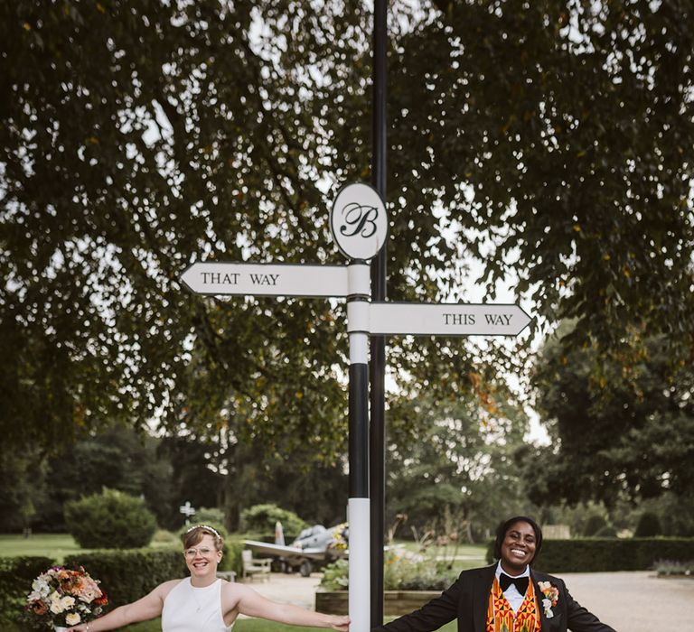 Bride in high neck sleeveless wedding dress holding on directional signage with bride in black suit jacket and Ghanian waistcoat 