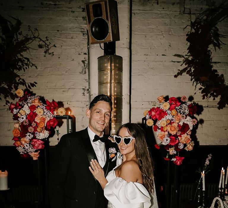 Bride in off the shoulder puff sleeve with groom in black tuxedo and colourful wedding flowers with sunglasses 