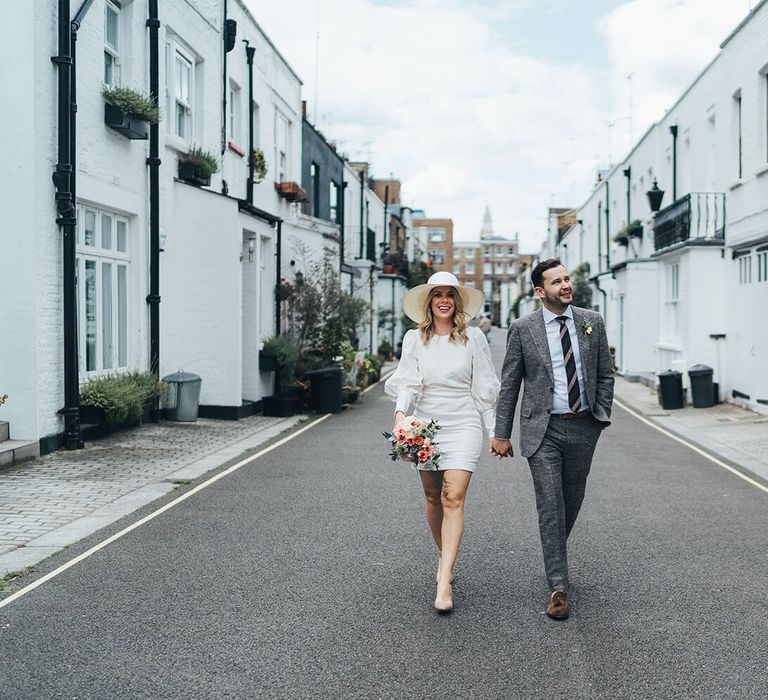 Bride and groom in London Marleybone holding hands room wears grey suit with brown and black striped tie, bride wears short wedding dress with puff sleeves, heels and a wedding hat