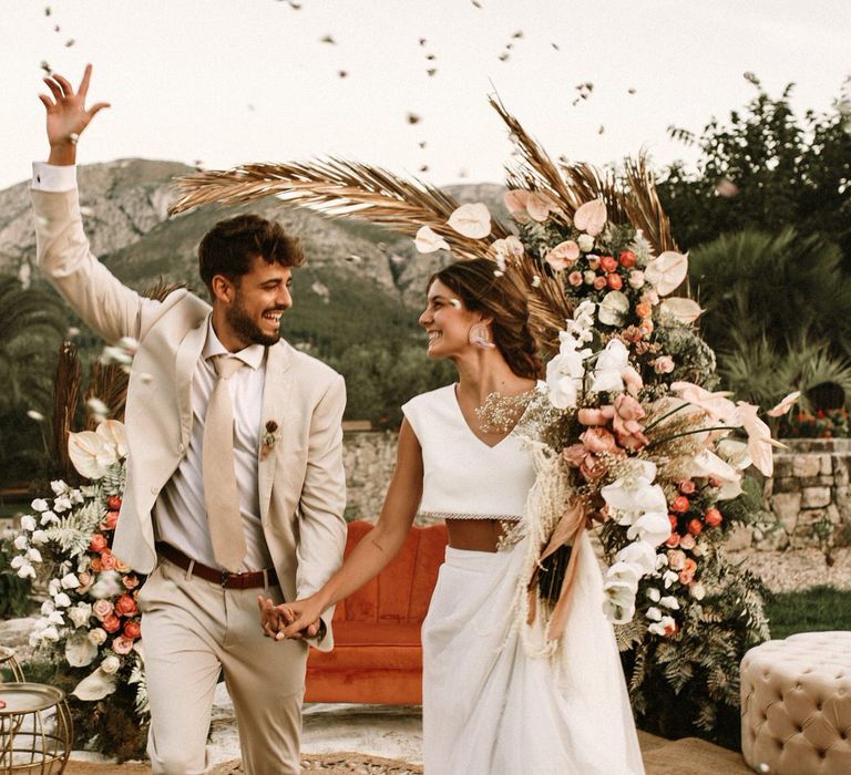 Confetti moment with boho bride in separates, groom in a beige suit with a boho wedding flowers arch 