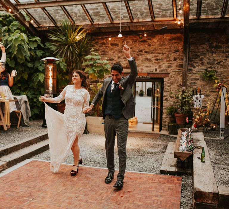 Bride in a lace Hermione de Paula wedding dress with front split entering the Anran glasshouse reception with her groom in a grey wool suit