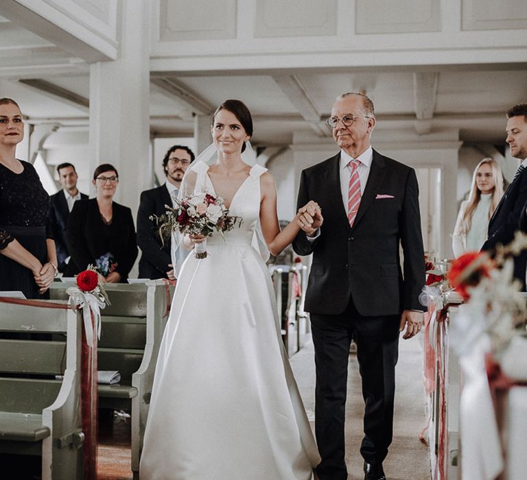 Smiling bride in white Jesus Piero wedding gown walks down the aisle with her father at German wedding