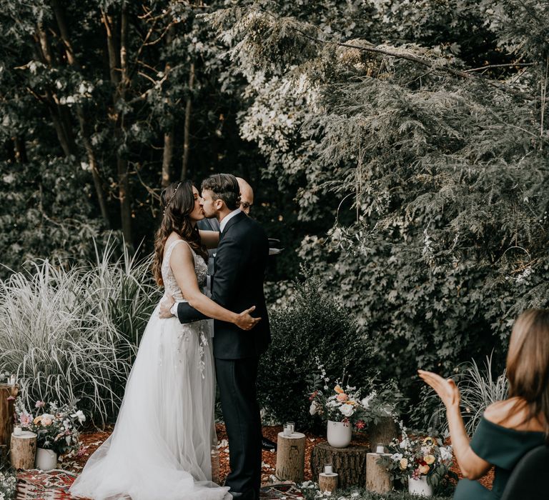 Bride & groom kiss during wedding ceremony surrounded by flowers and rustic scenery 