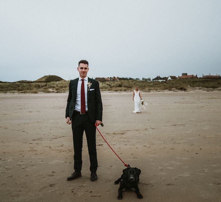 Groom stands with dog on the beach as he awaits first look moments