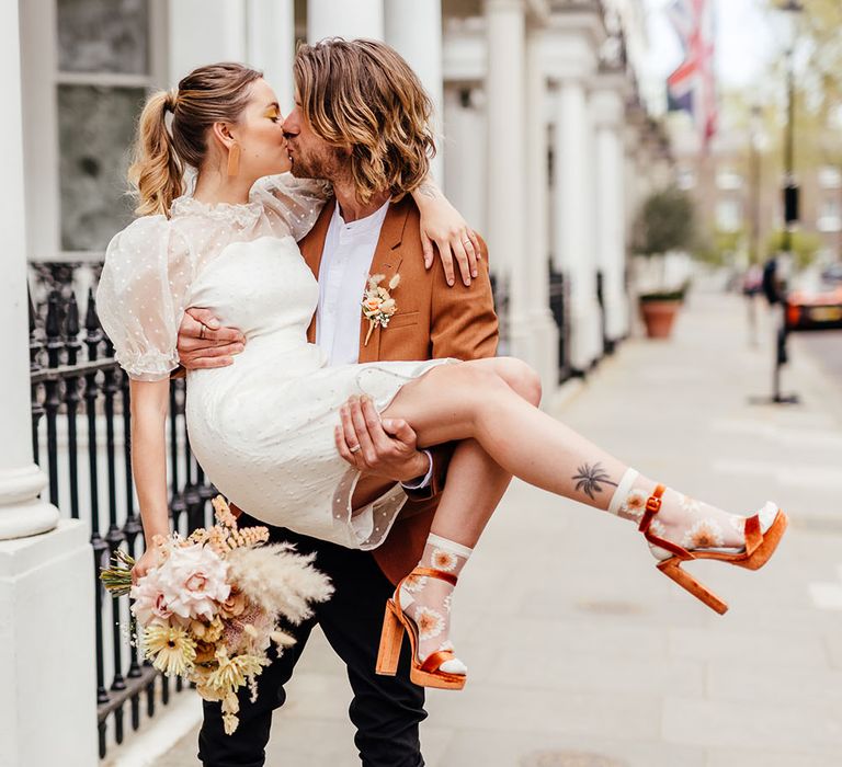 Groom in a rust coloured blazer picking his bride up in a polka dot short wedding dress and orange suede platform shoes 