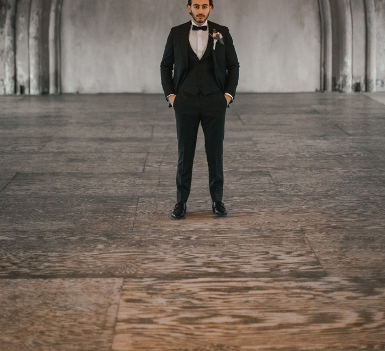 Groom stands in grey room whilst wearing black tie 