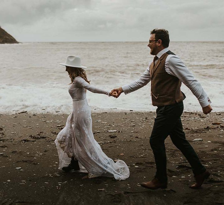 Bride in a long sleeve wedding dress, bridal boots and Fedora hat running on the beach with her husband 