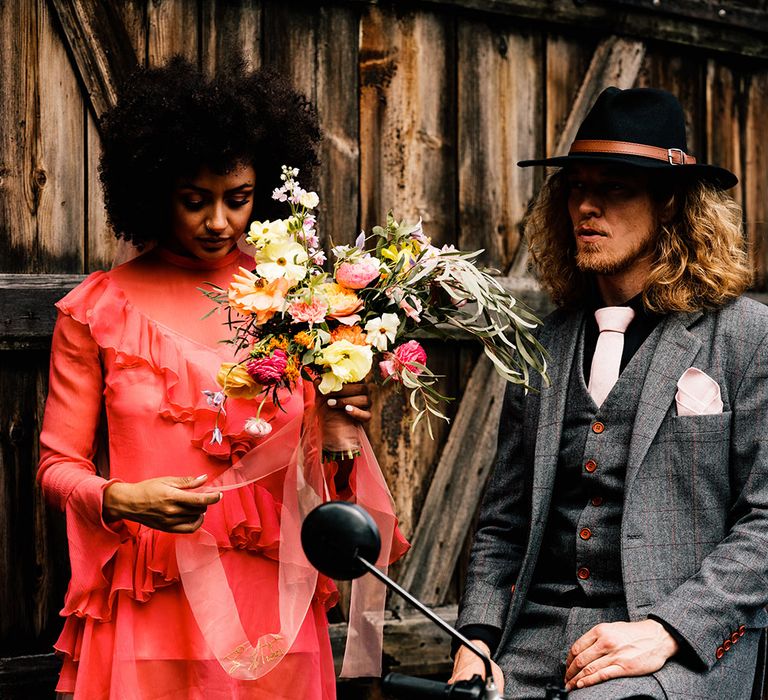 Bride in a red wedding dress with long sleeves holding a colourful wedding bouquet standing next to her groom in a grey check suit and black fedora hat 