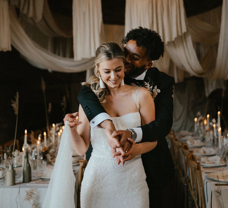 Groom in a tuxedo embracing his bride in a sparkly wedding dress as they stand inside their modern luxury wedding reception at Pentney Abbey, Norfolk