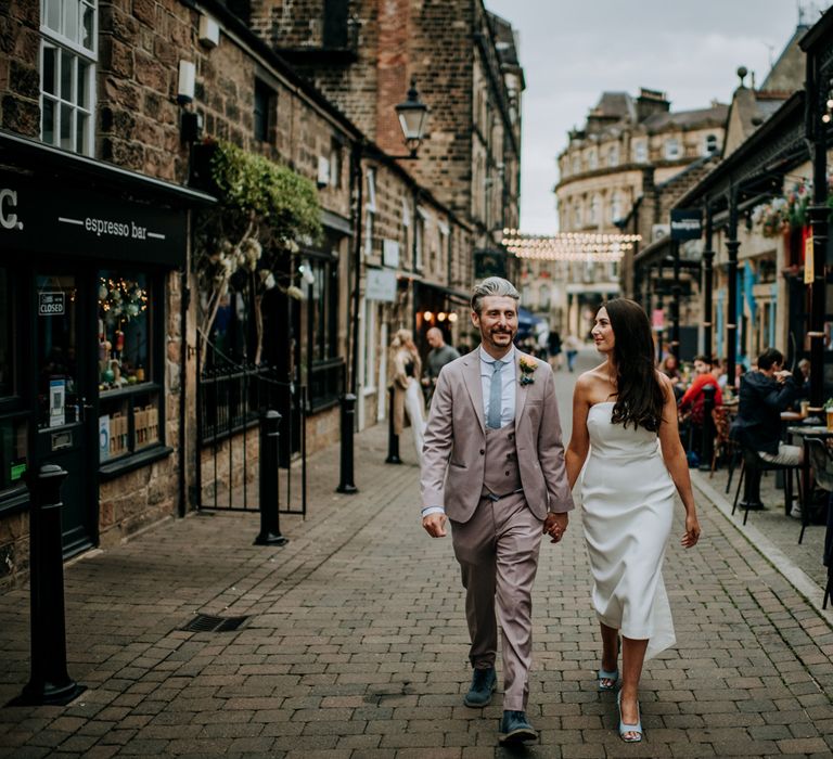 Bride in white strapless Rebecca Vallance Dress and blue slingback heels holds hands with groom in light brown Moss Bros suit and blue tie as they walk through streets of Harrogate after wedding