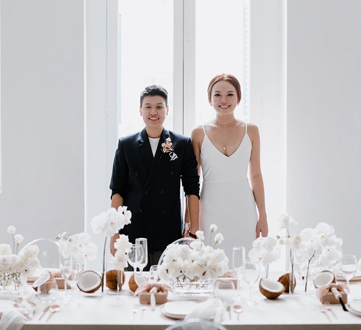 An Asian lesbian couple stand behind their wedding breakfast table in Singapore. One wears a suit jacket with sleeves rolled up and has cropped hair. tHe others wears a long with dress with hair tied loosely back.