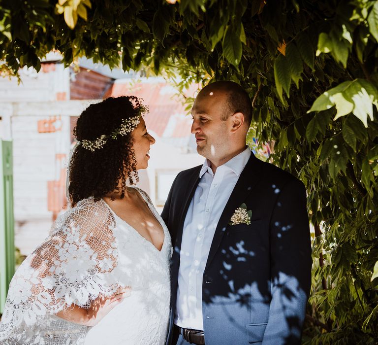 Bride & groom look lovingly at one another as they stand beneath greenery outdoors and the sun shines around them