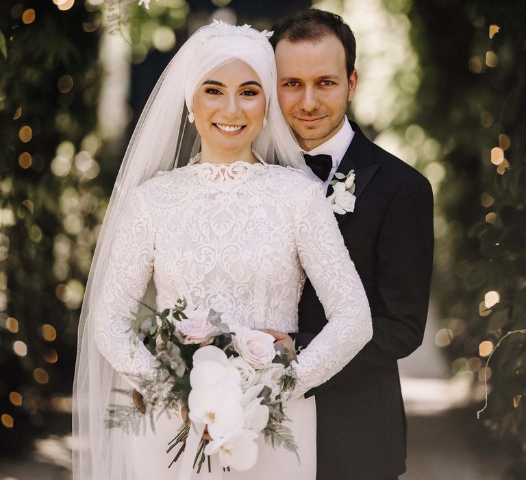 Groom embraces bride in long sleeve and high neck 3D lace wedding dress in matching veil under wisteria