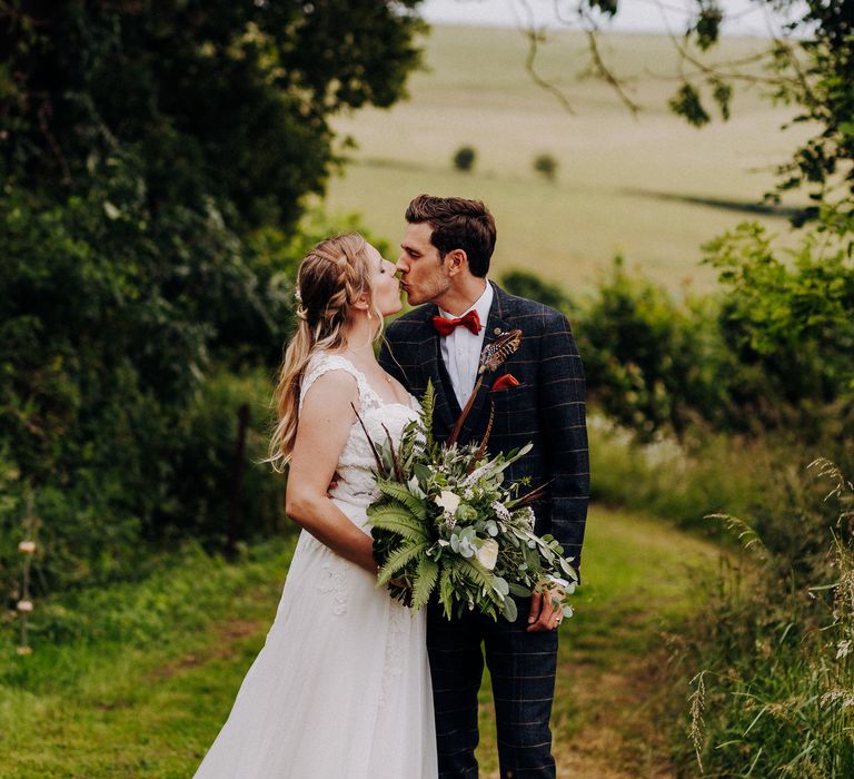 Bride & groom kiss one another in the countryside as groom wears Marc Darcy suit and bride wears Justin Alexander gown