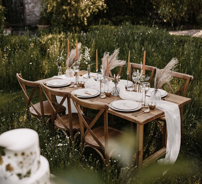 Wooden table and chairs in a meadow with dried flower and grasses in vases, gold candlesticks and orange taper candles 