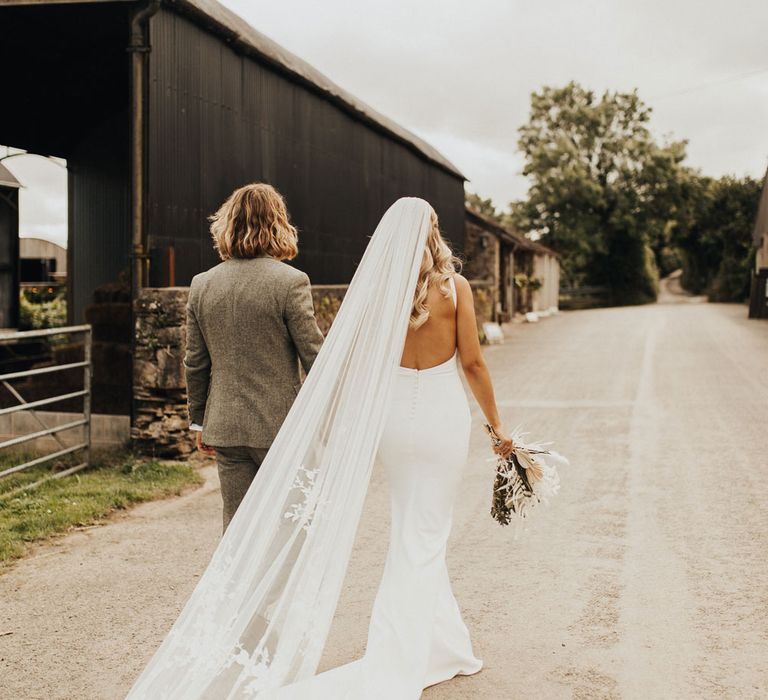 Bride in white open back Made With Love wedding dress with train and long embroidered veil holds white and green bridal bouquet and hand of groom in sage green suit in the other as they walk down country lane after wedding ceremony at Anran Devon