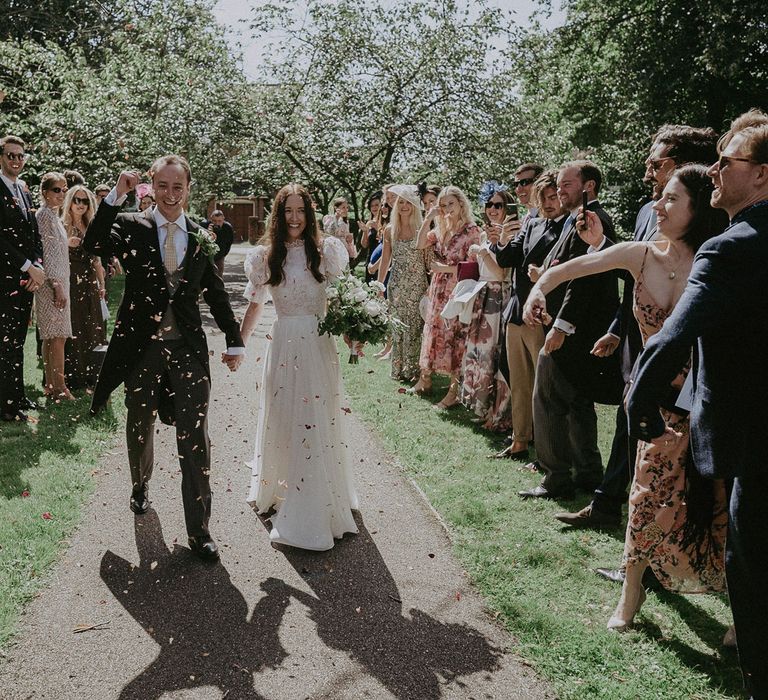 Smiling bride in white lace puffed sleeve Daalarna wedding dress holding white and green bridal bouquet walks down path holding hands with groom in black morning coat as guests throw confetti after church wedding ceremony in Surrey