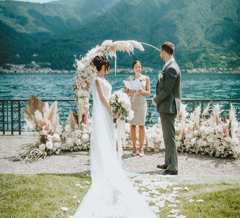 Bride & groom stand in front of floral installation during wedding ceremony with Lake Como in the background