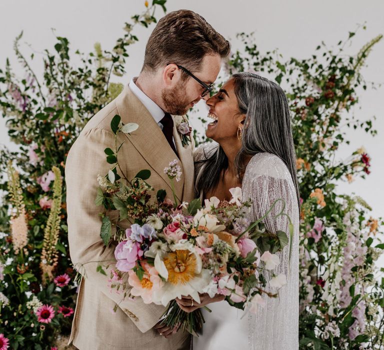 Bride in satin Halfpenny London wedding dress and tasselled bridal cape holding multicoloured bridal bouquet stands with groom in linen suit and gold Nike Air Force 1 trainers in front of floral installation for wedding at Loft Studios London