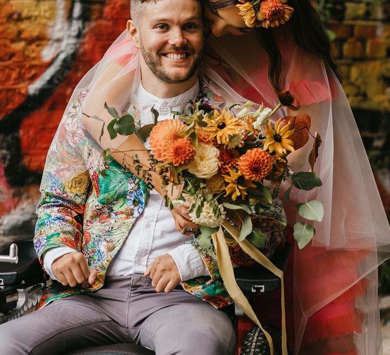 Bride in a coral wedding dress leaning over and embracing her C5/C6 Tetraplegic disabled groom in a patterned jacket 