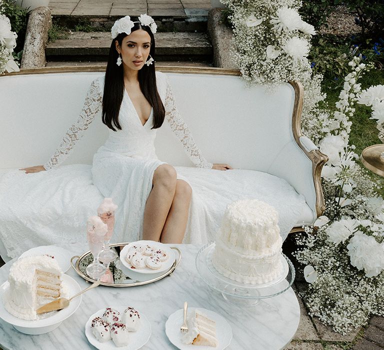 Tea party with white iced cake and biscuits and bride in a Constellation Ame lace wedding dress with long sleeves sitting on a vintage sofa 