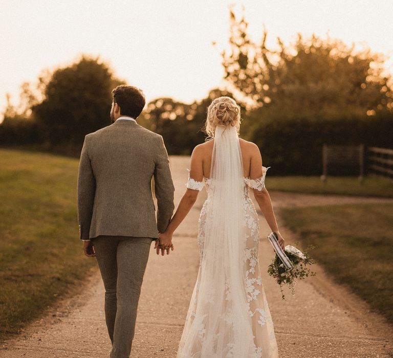 Bride in off the shoulder Enzoani wedding dress and veil holding mixed bridal bouquet walks down country lane holding hands with groom in grey suit at golden hour during Inkersall Grange Farm wedding