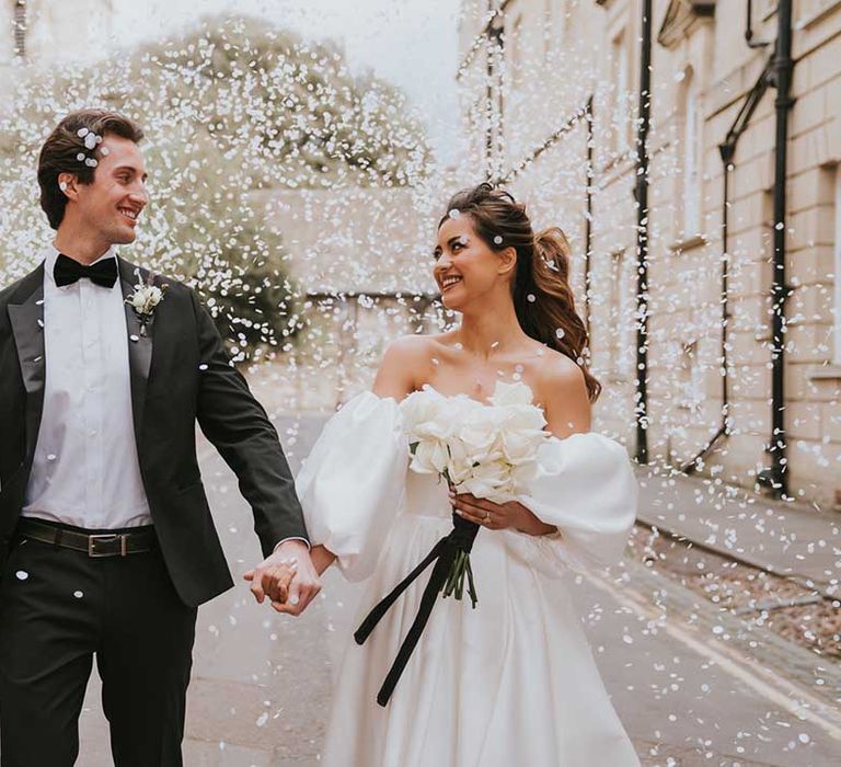 White confetti moment at black tie wedding with groom in a tuxedo and bride in a strapless wedding dress holding a white rose wedding bouquet 