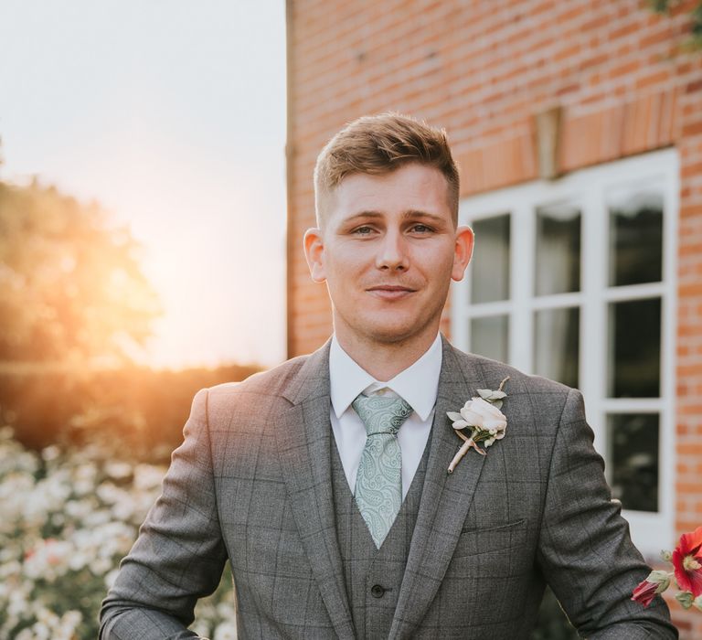 Groom in grey checked three piece suit, mint green paisley tie and floral button hole buttons up suit as he stands in garden at Primrose Hill Farm during golden hour for summer wedding