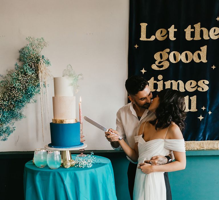 Groom in an open collar white shirt kissing his bride in a cold shoulder wedding dress as they cut their contemporary wedding cake 