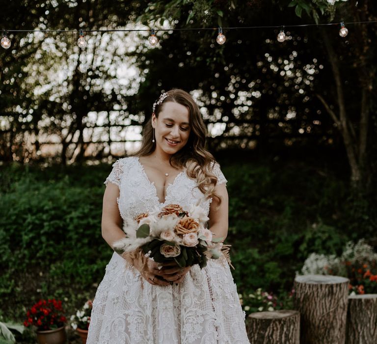 Plus size bride in homemade lace wedding dress with train holding mixed rose bridal bouquet stands in garden after wedding ceremony