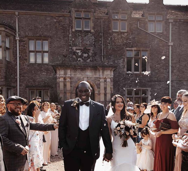 Groom wears dark suit with a white shirt & bow tie holding hands with Bride in long half chiffon detail dress at Cefn Tilla Court as their guests celebrate 