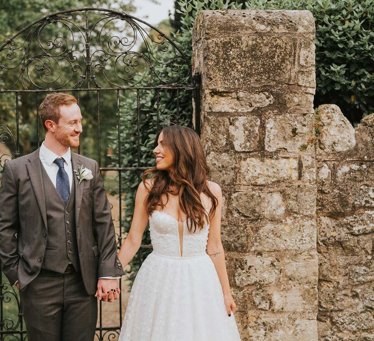 Groom in grey three piece suit, blue tie and white rose buttonhole smiles whilst holding hands with bride in cami strap Pronovias wedding dress as they stand by walled garden in Buckinghamshire