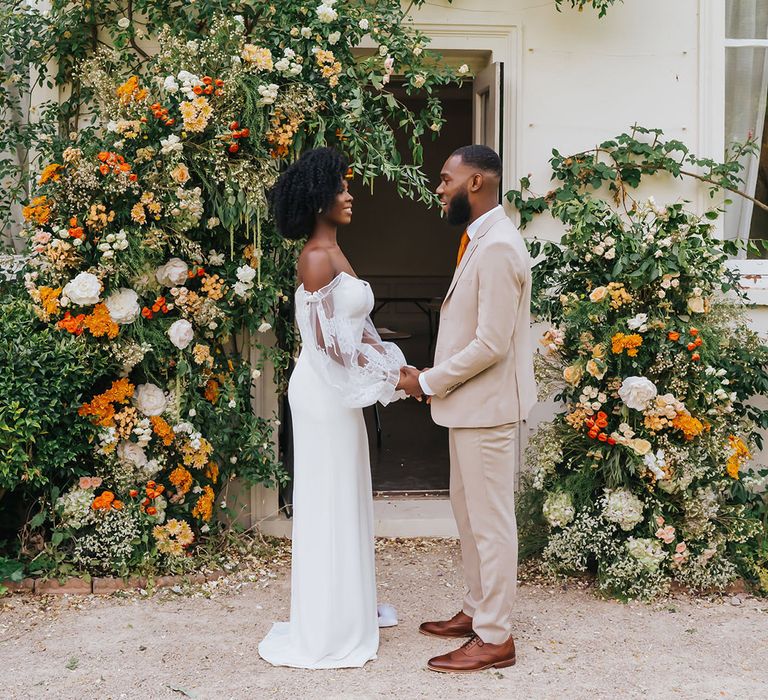 Bride in a strapless wedding dress with sheer sleeves and groom in beige suit holding hands next to the vine at Modern Hall, London with orange flowers